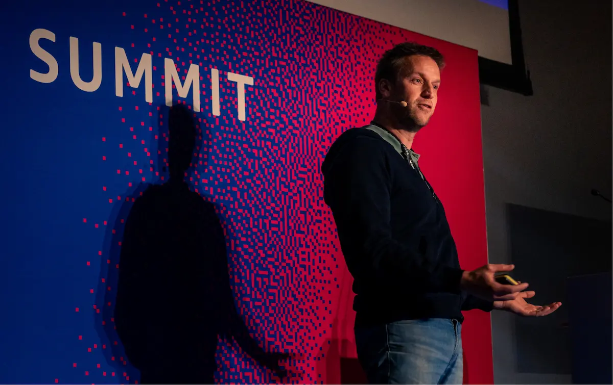 Speaker Tom Jansen presenting on stage at AWS Summit Amsterdam with blue and red digital pattern backdrop.